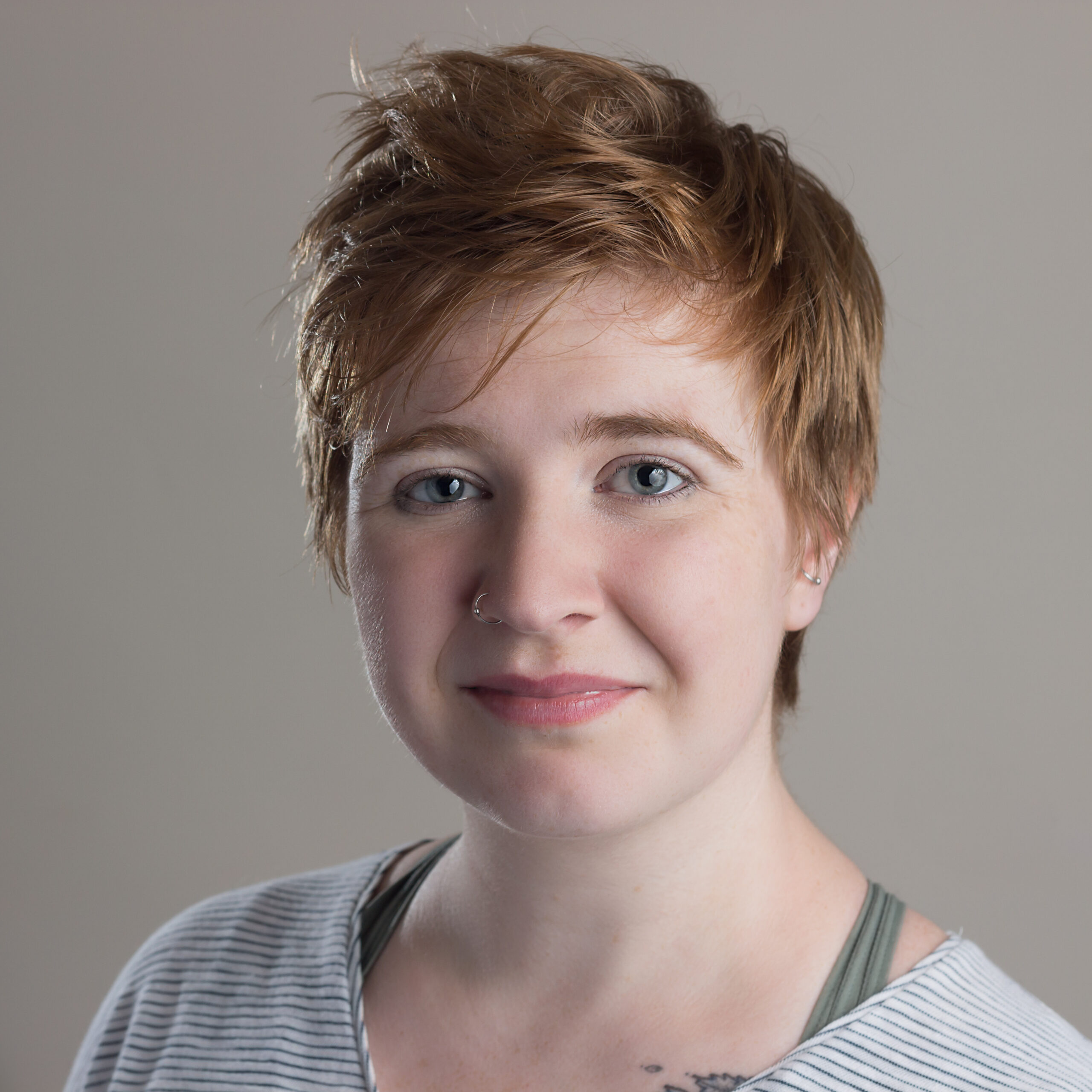 A young person with short ginger-brown hair and blue eyes, standing in front of a neutral studio background.
