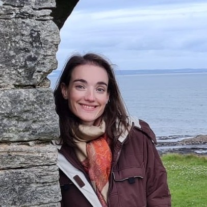 A young woman with long dark hair, wearing a scarf and raincoat, leaning against a stone wall in front of a body of water.