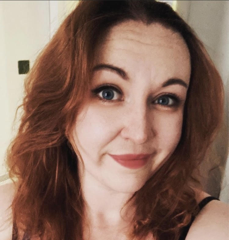 A young woman with wavy red hair and red lipstick smiling against a white background.