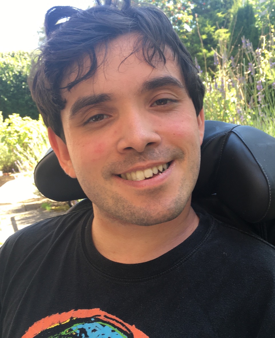 A young man with short dark hair and a printed tshirt, smiling and sitting in a wheelchair against a leafy background.