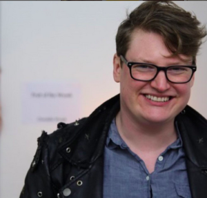 A young person with short brown hair and dark-rimmed glasses, wearing a button-up denim shirt and smiling.