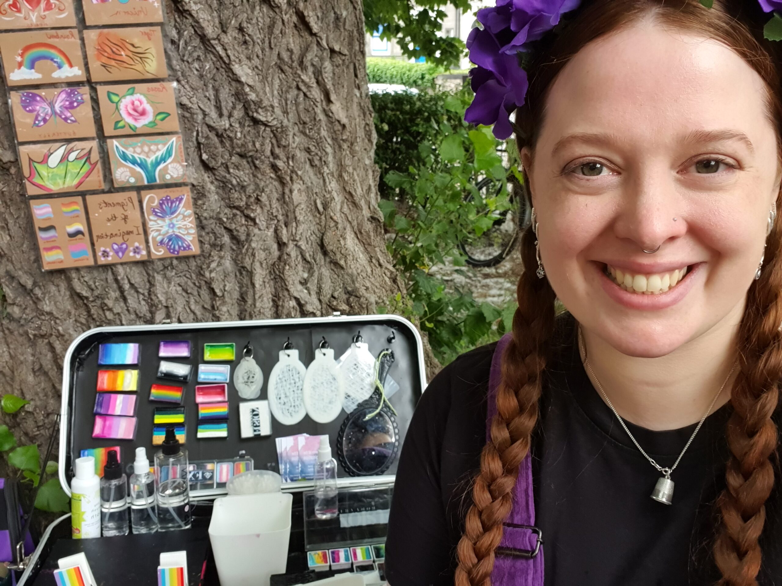 A young woman with long braids and a purple hairpiece, wearing a silver necklace and a dark top, standing against a leafy background and smiling.