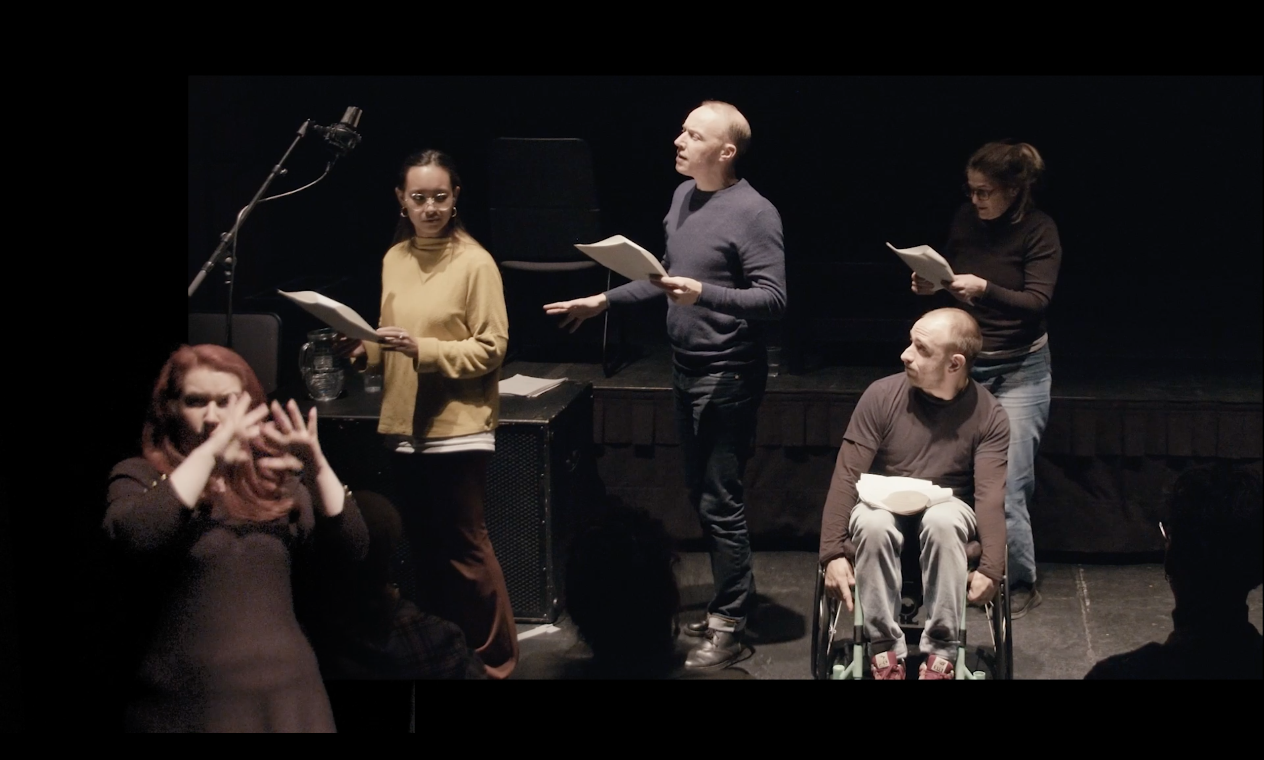 Four actors performing in a script-in-hand reading. A BSL interpreter is overlaid in the bottom left corner.