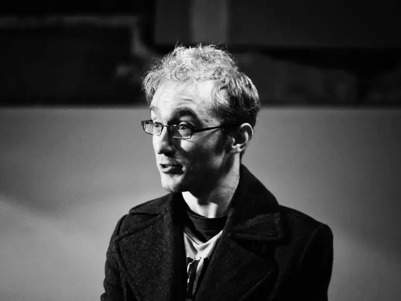 A young man with short hair, wearing glasses and a black cardigan. He is harshly lit and looking to one side, in the middle of saying something.