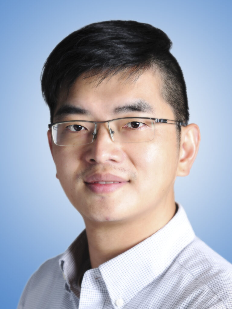 A young East Asian man with glasses and a neutral expression. He is wearing a business shirt and posed against a blue backdrop.