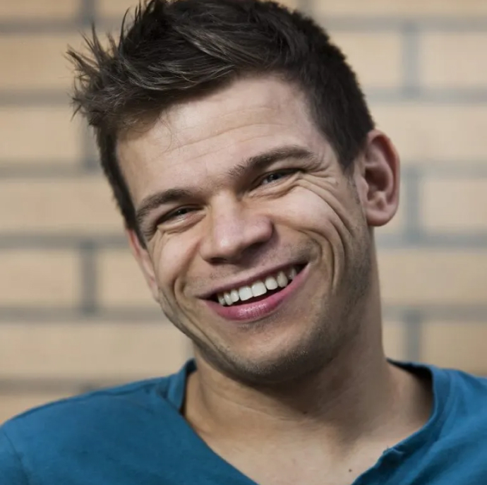 Robert is smiling at the camera. He is a white man with short dark hair. He's wearing a blue t-shirt and has a brick background behind him.